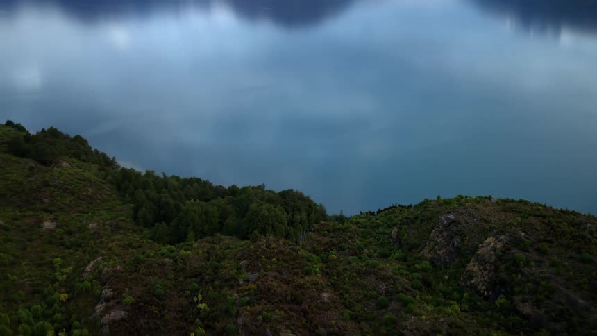 An aerial pullback over a lush hillside reveals a vast, serene mountain lake reflecting the cloudy sky and distant snow-capped peaks