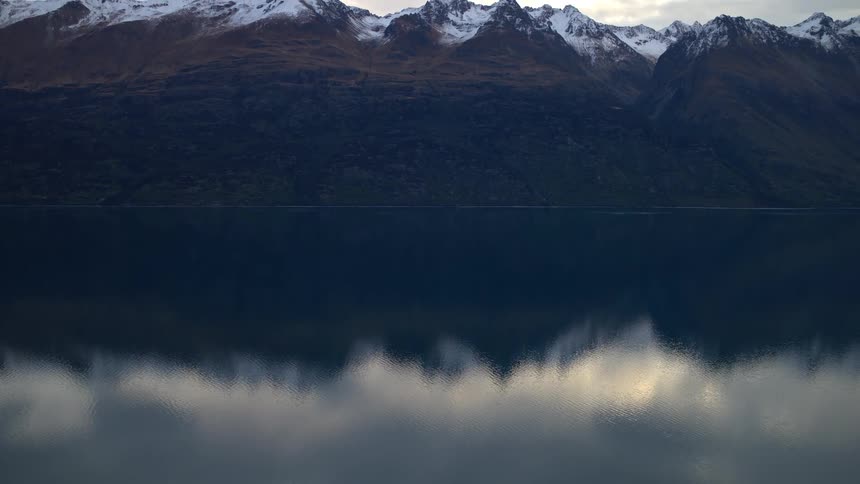 A slow zoom out reveals a majestic, snow-capped mountain range perfectly reflected in the still waters of a serene lake at twilight