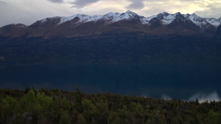 Slow aerial pullback over a windy forest reveals a serene lake reflecting snow-capped mountains at dusk