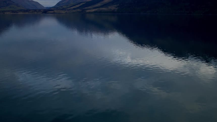 A slow tilt-up from the dark, rippling waters of a lake reveals a stunning mountain landscape with snow-capped peaks under a cloudy sky