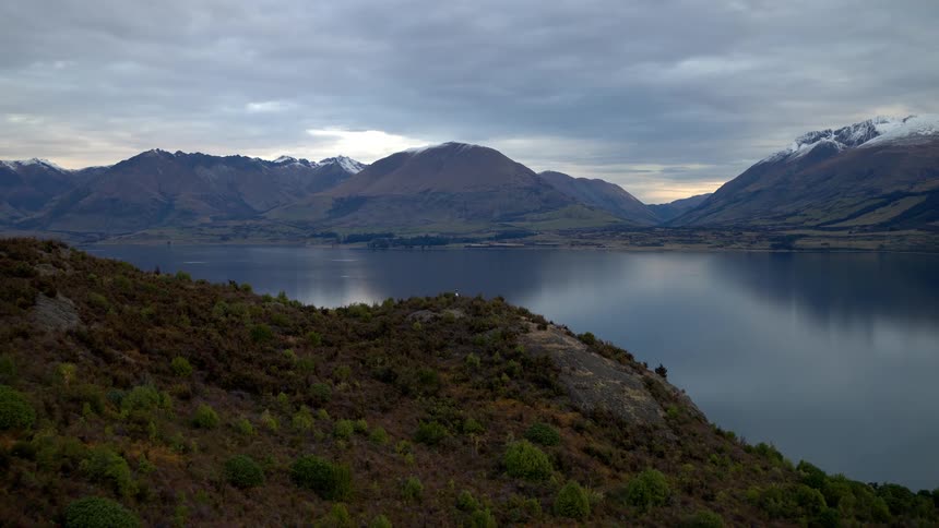 An expansive aerial shot slowly pushes over a brush-covered hillside to reveal a serene lake reflecting snow-capped mountains under a cloudy sky