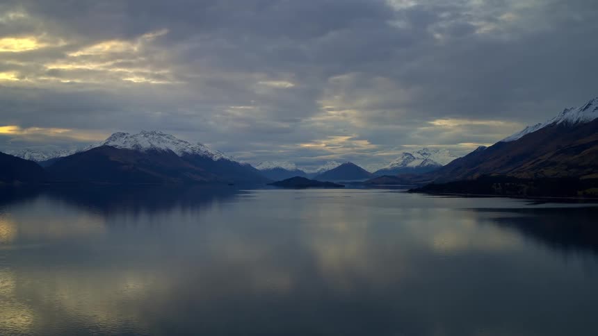 A slow aerial pullback reveals a vast, serene lake reflecting snow-capped mountains under a moody, dramatic sky