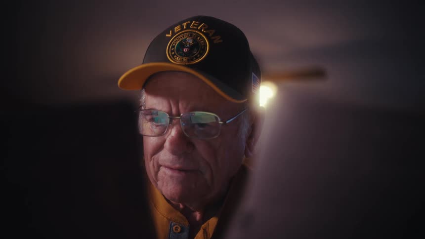 A close-up of an elderly veteran smiling and then becoming serious as he reads from a brightly lit screen in a dark room