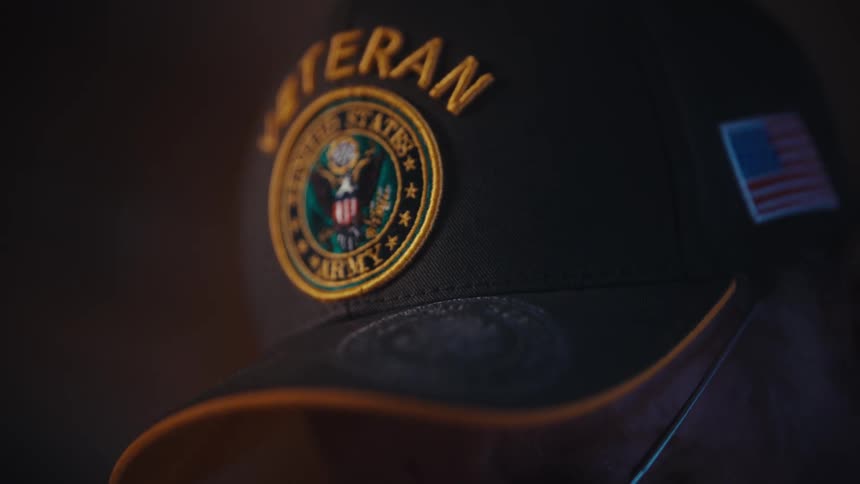 A somber elderly US Army veteran wearing a 'Veteran' hat is shown in a dimly lit, thoughtful close-up