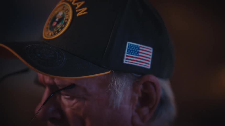 A contemplative elderly United States Army veteran slowly puts on his glasses in a dimly lit, intimate setting