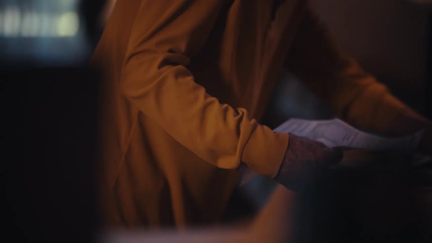 An elderly person's wrinkled hands sort through a pile of financial paperwork and bills at a table in a dimly lit room