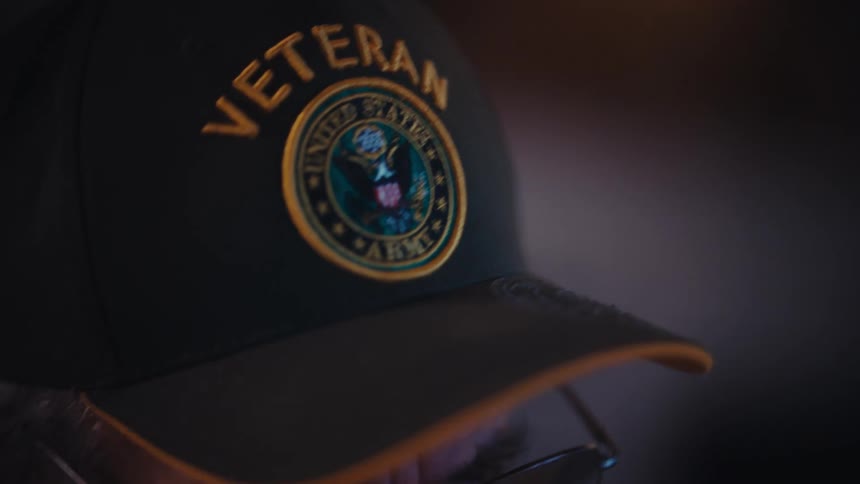 Close-up of a contemplative elderly Army veteran wearing a hat and glasses as he looks down in a dimly lit room
