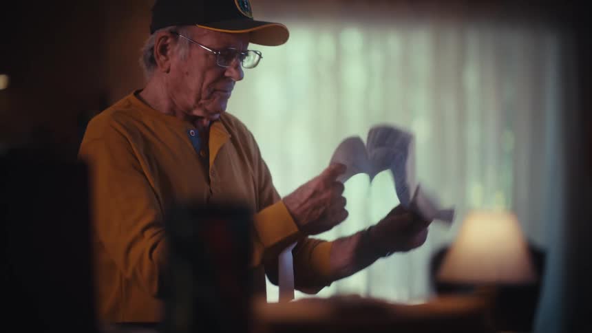 An elderly veteran with a concerned expression is overwhelmed by a long bill while reviewing his expenses at home