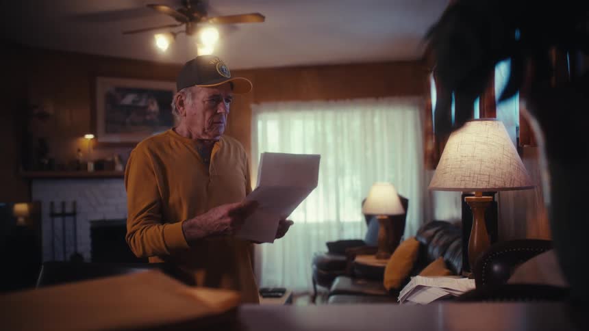 A worried elderly man in a baseball cap stands in his living room and reads over bills in slow motion