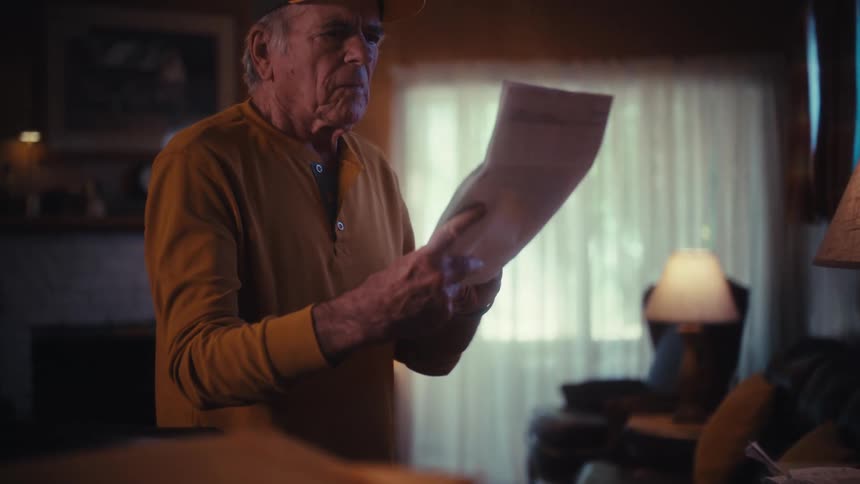 An elderly man in a baseball cap stands in his living room, carefully reading a document with a serious expression