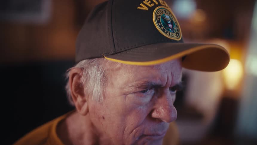 A worried elderly US Army veteran wearing a hat holds a financial statement with interest charges in a dimly lit home