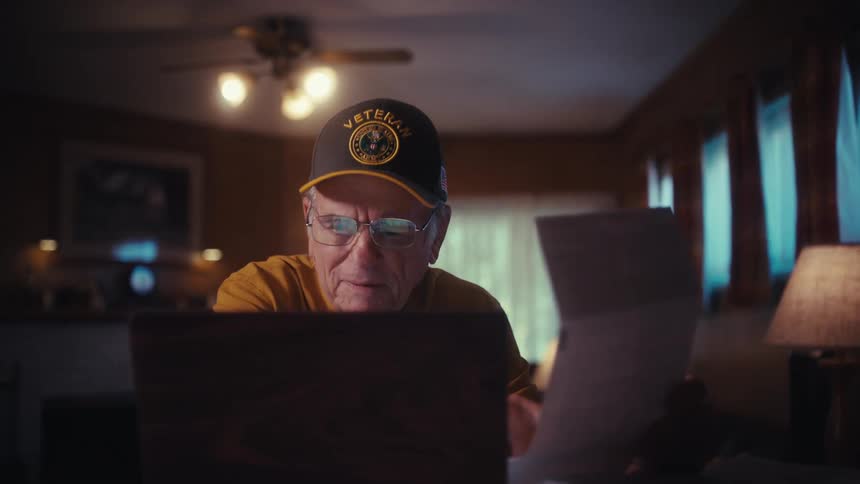 A happy elderly veteran wearing a cap smiles with relief as he manages his finances online using a laptop at his desk at home