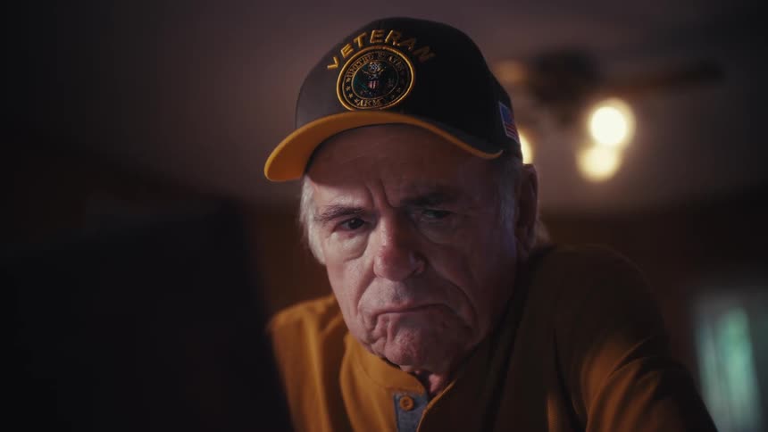 An elderly military veteran wearing a hat looks at a screen with a sad and worried expression in a dimly lit room