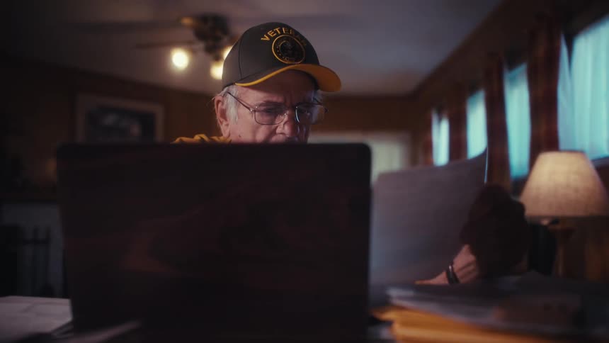 Slow zoom shot of an elderly veteran concentrating as he reviews paperwork while using his laptop at home