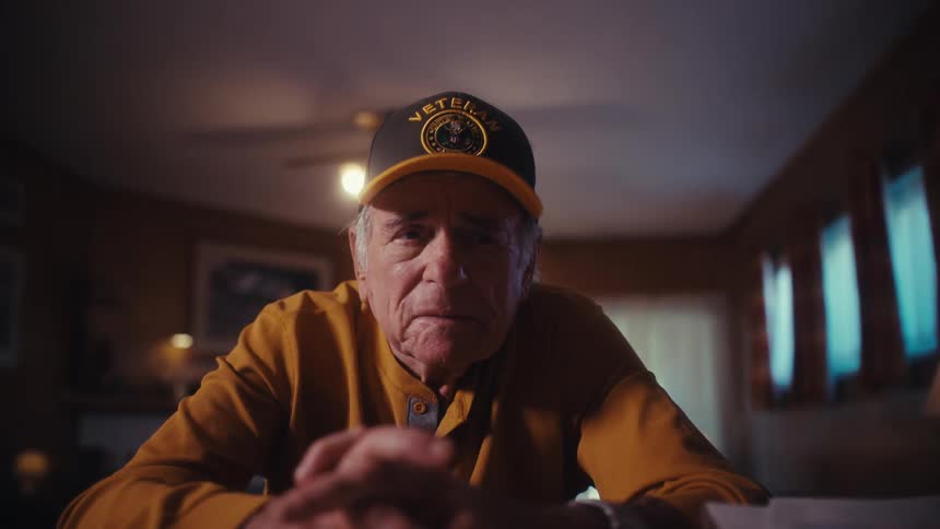 A somber elderly veteran with a serious expression sits alone at his desk in a dimly lit room