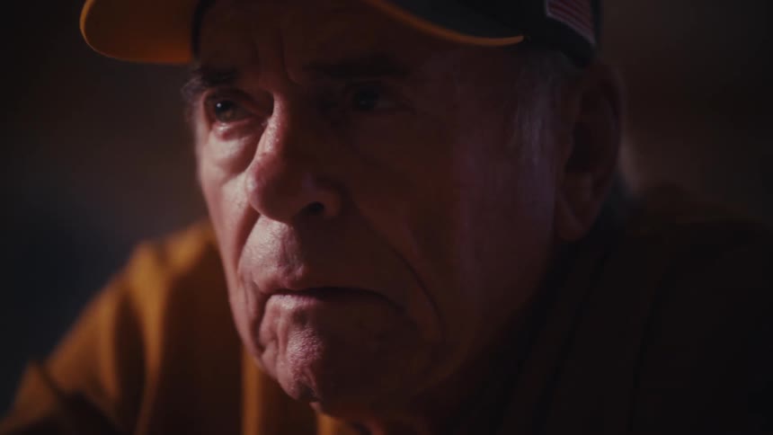 A close-up of a solemn elderly U.S. Army veteran in a dark room, reflecting with a pensive expression