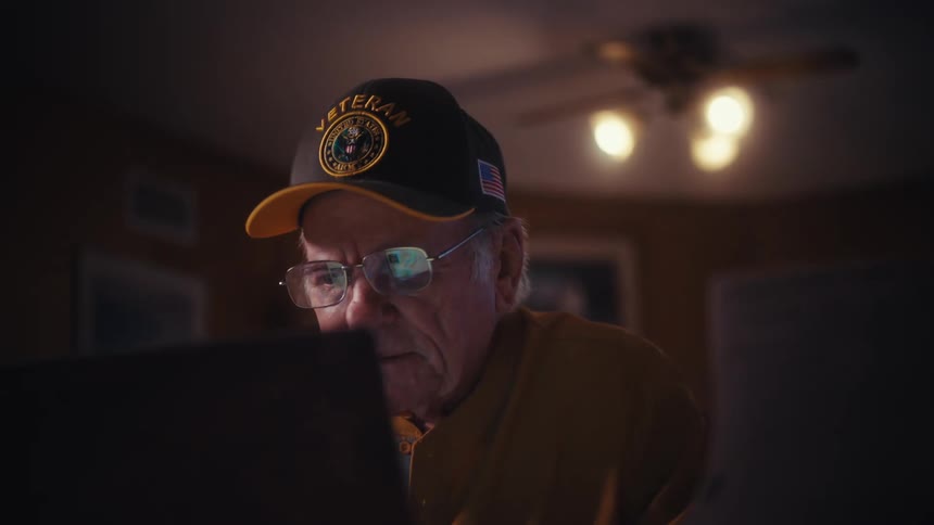 A senior veteran sits at his desk reading a letter, then smiles happily while using his laptop in his cozy home