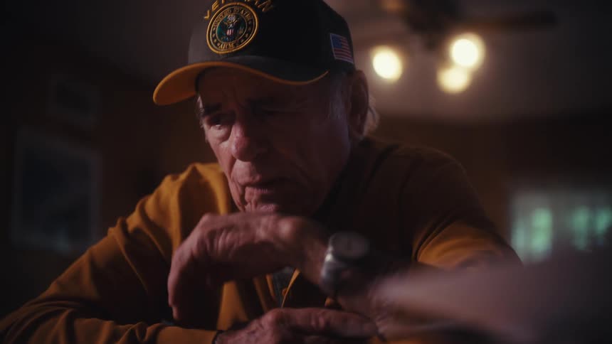 An elderly veteran wearing a cap slowly looks up with a sad, contemplative expression in a dimly lit room