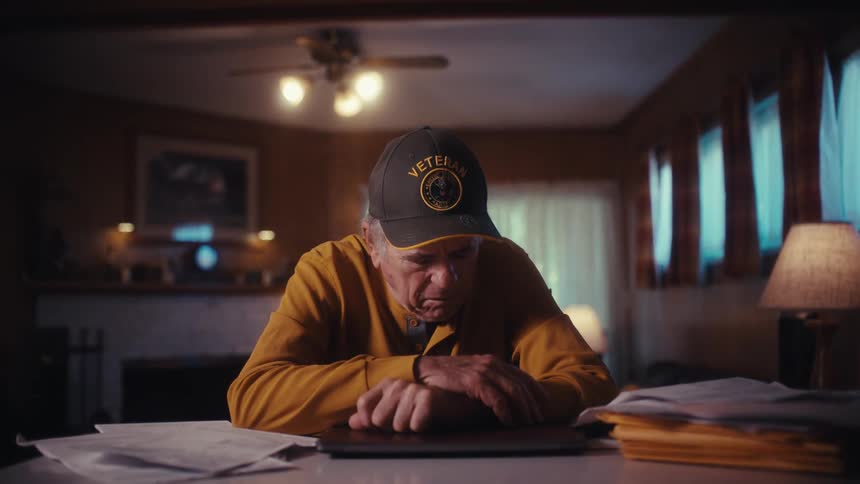 A stressed elderly veteran wearing a cap closes his laptop and bows his head in defeat after reviewing his bills at home