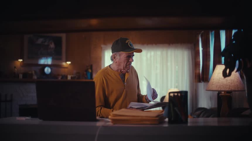 A solemn elderly veteran carefully reads through his bills and paperwork while sitting at his desk at home