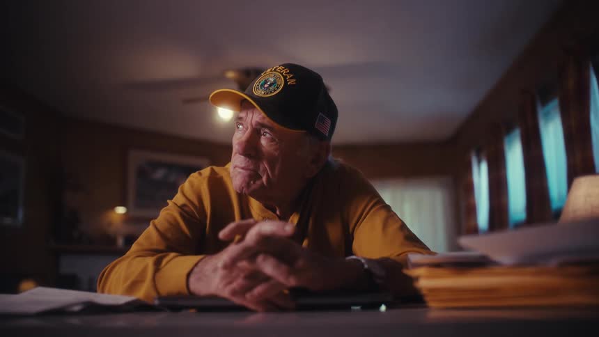 A thoughtful elderly veteran sits alone at a table in his home, looking off into the distance