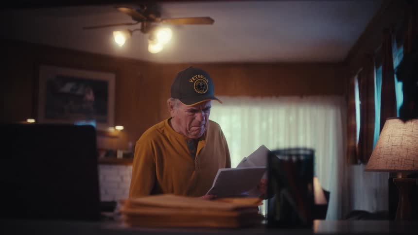 A serious elderly veteran in a dimly lit home office carefully reviews important documents and bills from his mail