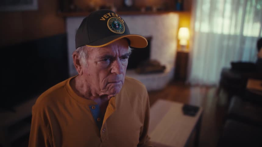 An elderly veteran wearing a cap takes off his glasses and looks at a document with a worried expression in his dimly lit living room