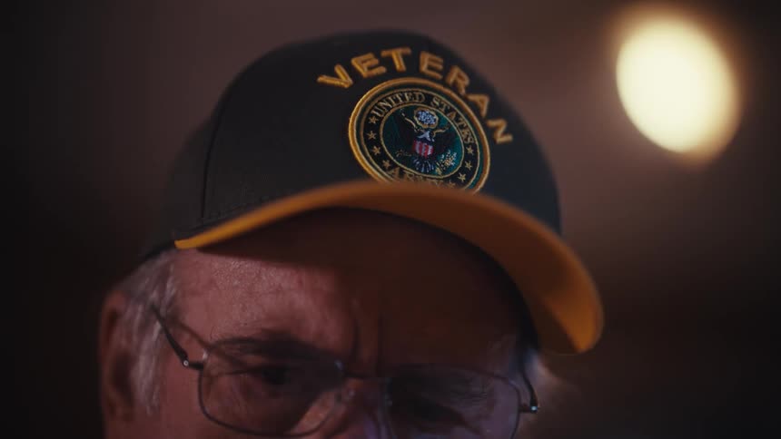 A solemn elderly man wearing a United States Army veteran hat sits in a dimly lit room