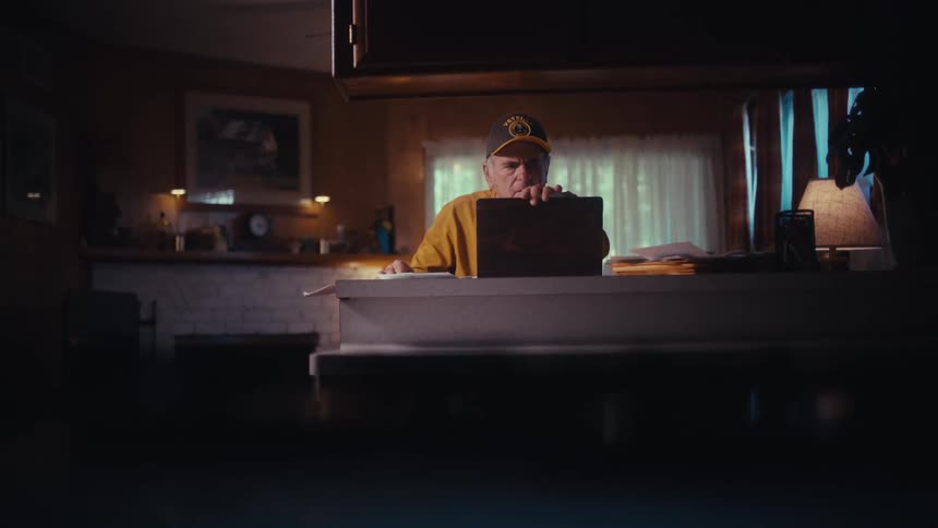 A distraught elderly man in a hat closes his laptop and sits alone at his kitchen counter, lost in worried thought