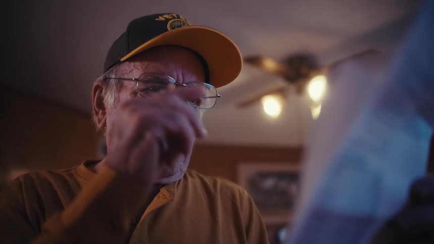 A worried elderly veteran with a stressed expression removes his glasses as he struggles to comprehend a shocking bill