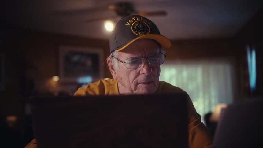 An elderly veteran sits at his desk in a dimly lit room, looking over paperwork and bills while using his laptop to manage finances