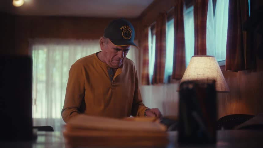 A serious elderly veteran wearing a hat stands at a table in his dimly lit home, carefully reviewing his bills and paperwork