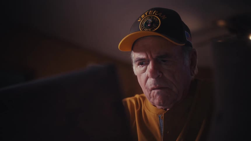 A pensive elderly veteran stares intently at a laptop screen in a dimly lit room, reflecting on what he sees