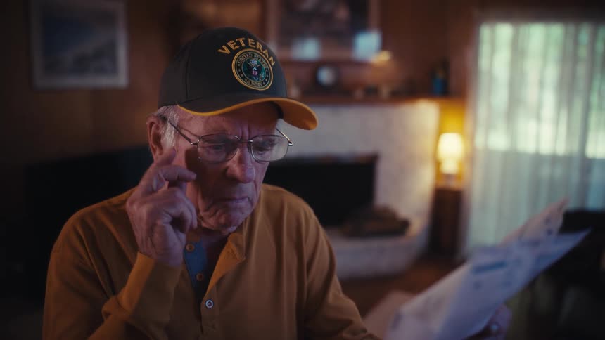 An elderly veteran in a cap looks stressed and worried while reviewing a bill or document in his home