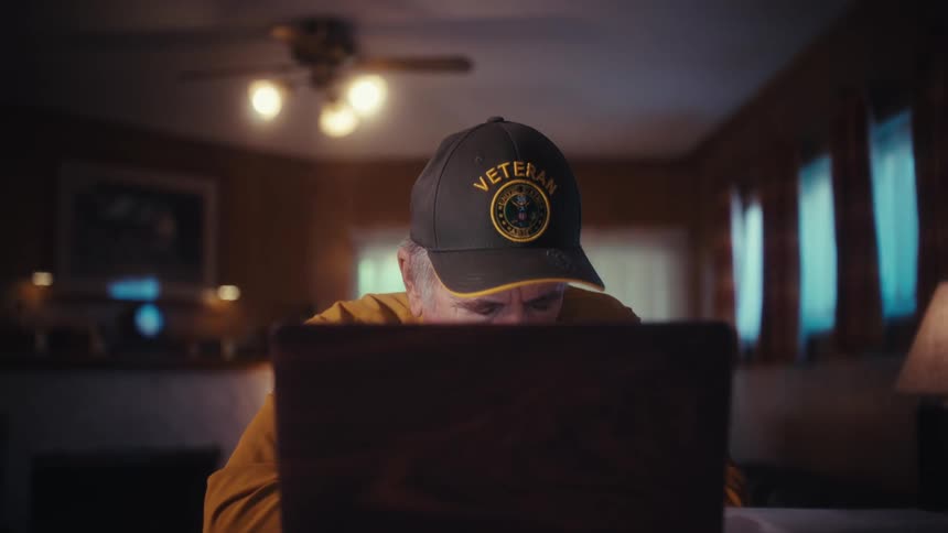 A cinematic slow-motion dolly shot shows an elderly veteran looking stressed while reviewing documents and working on his laptop at home