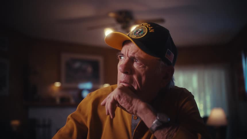 A worried elderly veteran closes his laptop and contemplates his financial problems while sitting at his desk in a dimly lit home
