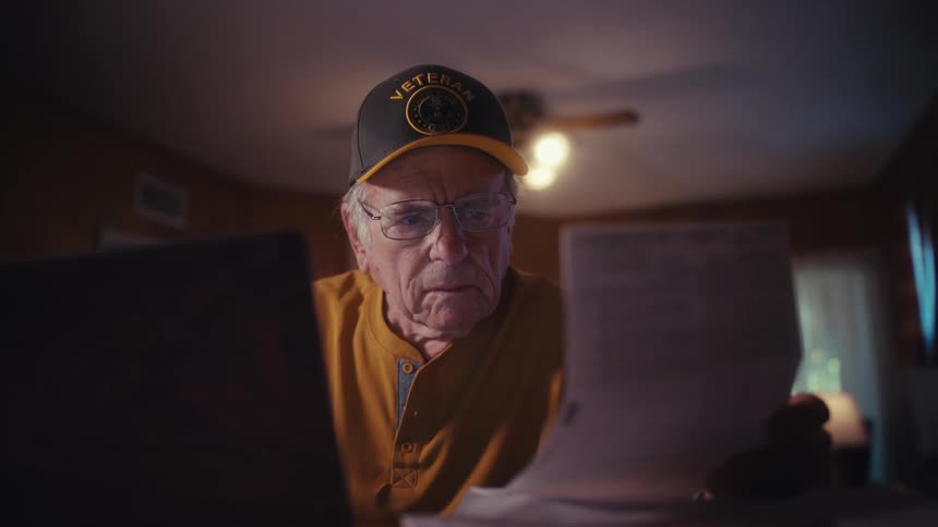 An elderly veteran with a concerned expression uses his laptop to manage his finances and pay bills at home