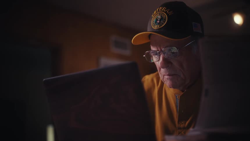 Elderly veteran carefully reviews documents and manages his affairs on a laptop at his desk late at night