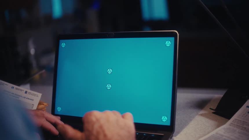 An older man types on a laptop computer with a green screen at a desk in a dimly lit room at night