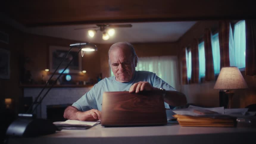 An elderly Army veteran at his desk puts away paperwork, opens his laptop, and smiles with happy surprise