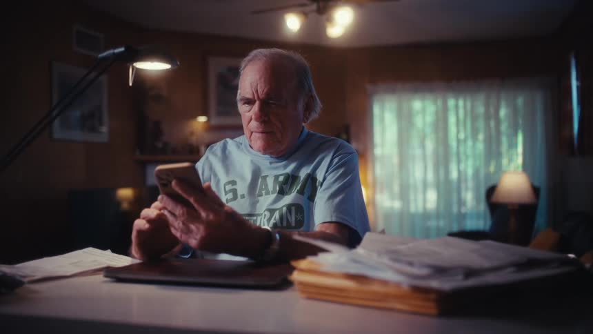 A serious elderly U.S. Army veteran sits at a desk in his dimly lit home, concentrating on his smartphone