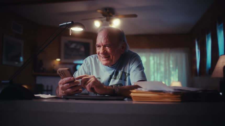 A joyful senior man laughs heartily while scrolling on his smartphone at a desk in his home