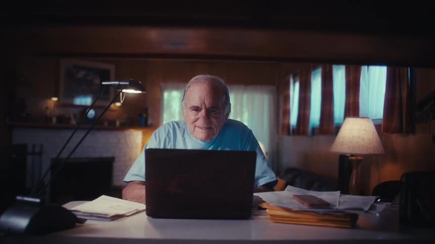 A slow push-in shot of an elderly US Army veteran opening his laptop computer at his desk in his dimly lit home