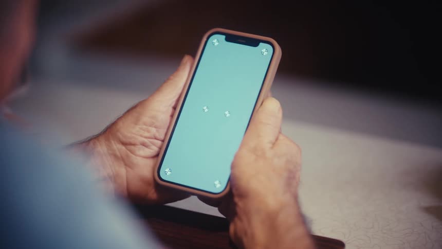An over-the-shoulder view shows an older man holding a smartphone with a blank green screen perfect for custom content