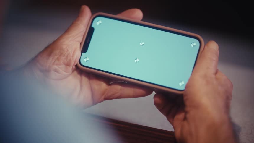 An over-the-shoulder view of an elderly person holding a smartphone horizontally with a green screen ready for compositing