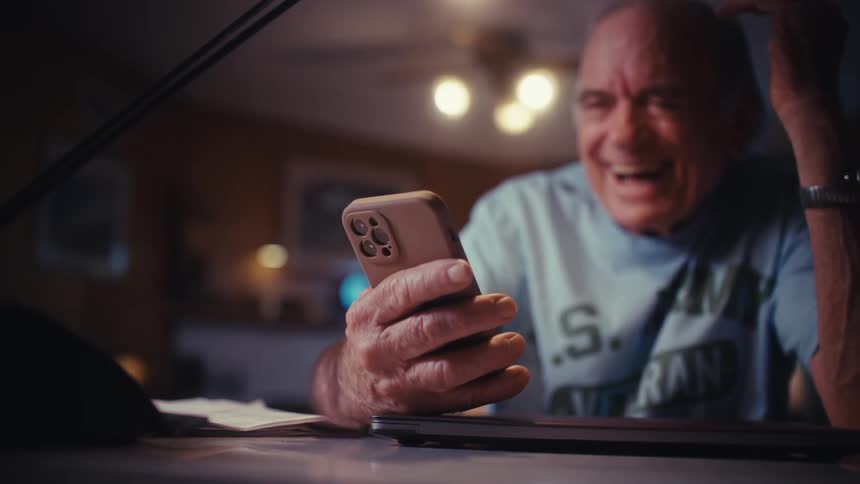 A happy senior veteran enjoys a video chat on his phone, laughing with delight at his desk in the evening