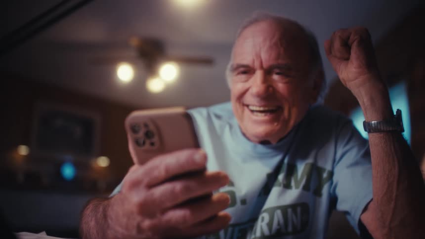A joyful elderly Army veteran celebrates while looking at his smartphone, laughing and pumping his fist in the air with excitement