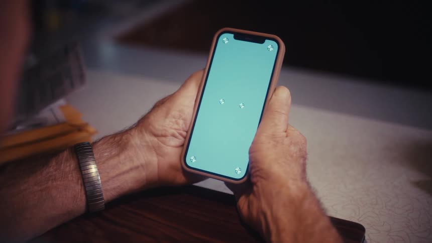 An over-the-shoulder shot shows an elderly man's hands holding a smartphone with a green screen