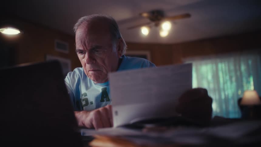 A stressed elderly Army veteran struggles with his finances while reviewing bills on his laptop late at night in his home