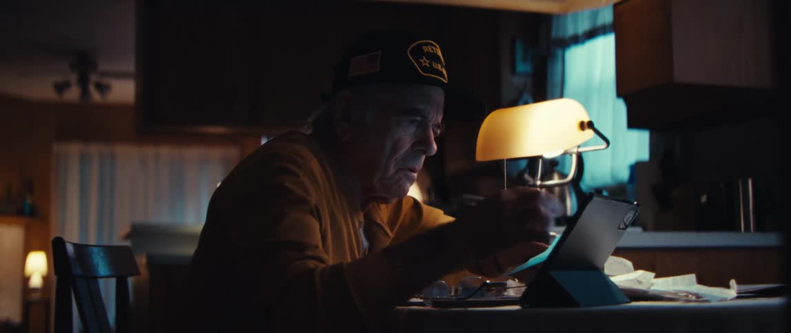 An elderly veteran in a hat sits at a dimly lit table at night, using a tablet and stylus to manage his finances